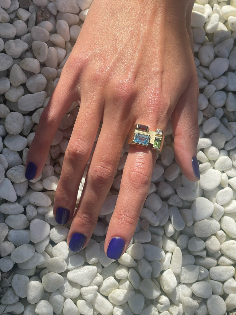 A hand wearing a gold ring with a baguette cut garnet, peridot, and sky blue topaz on a pebble surface.